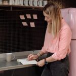 Woman standing beside a pink retro refrigerator taking notes in a kitchen